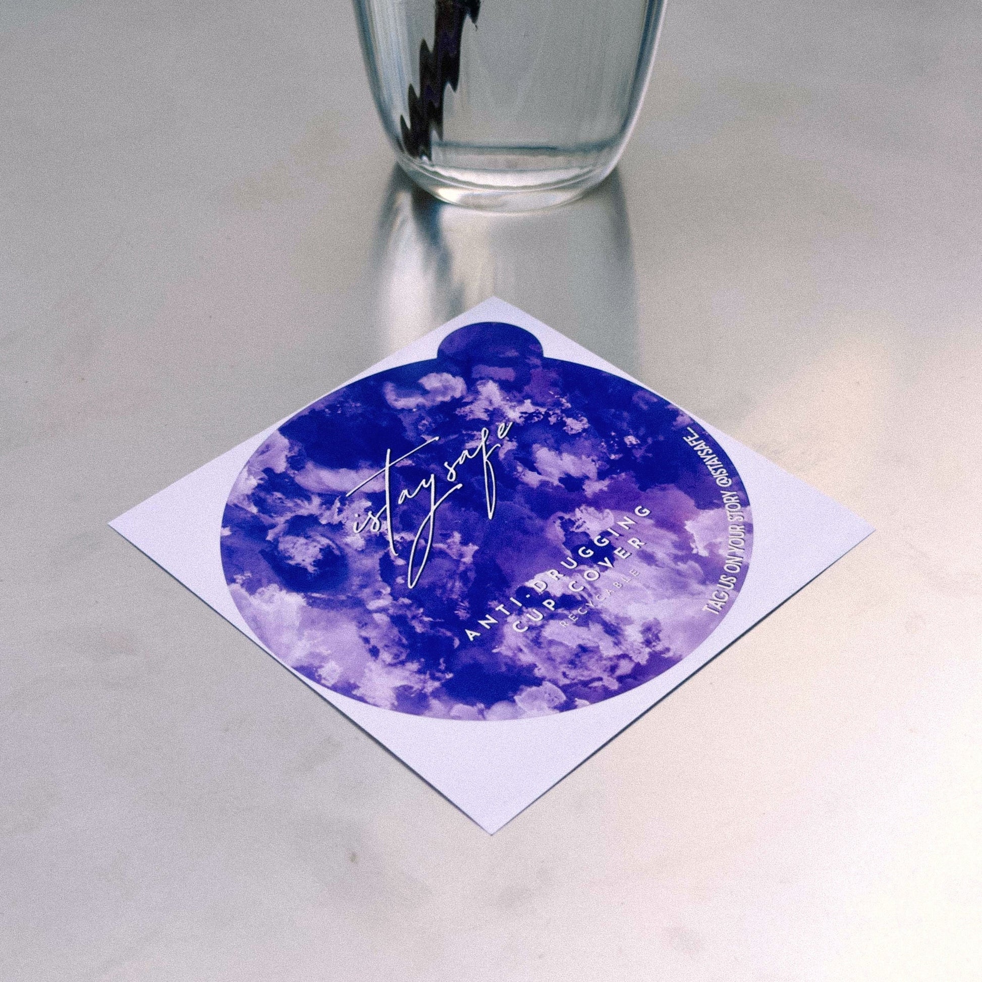 Purple ISTAYSAFE cup cover laying on the table, with a glass of water in the background.