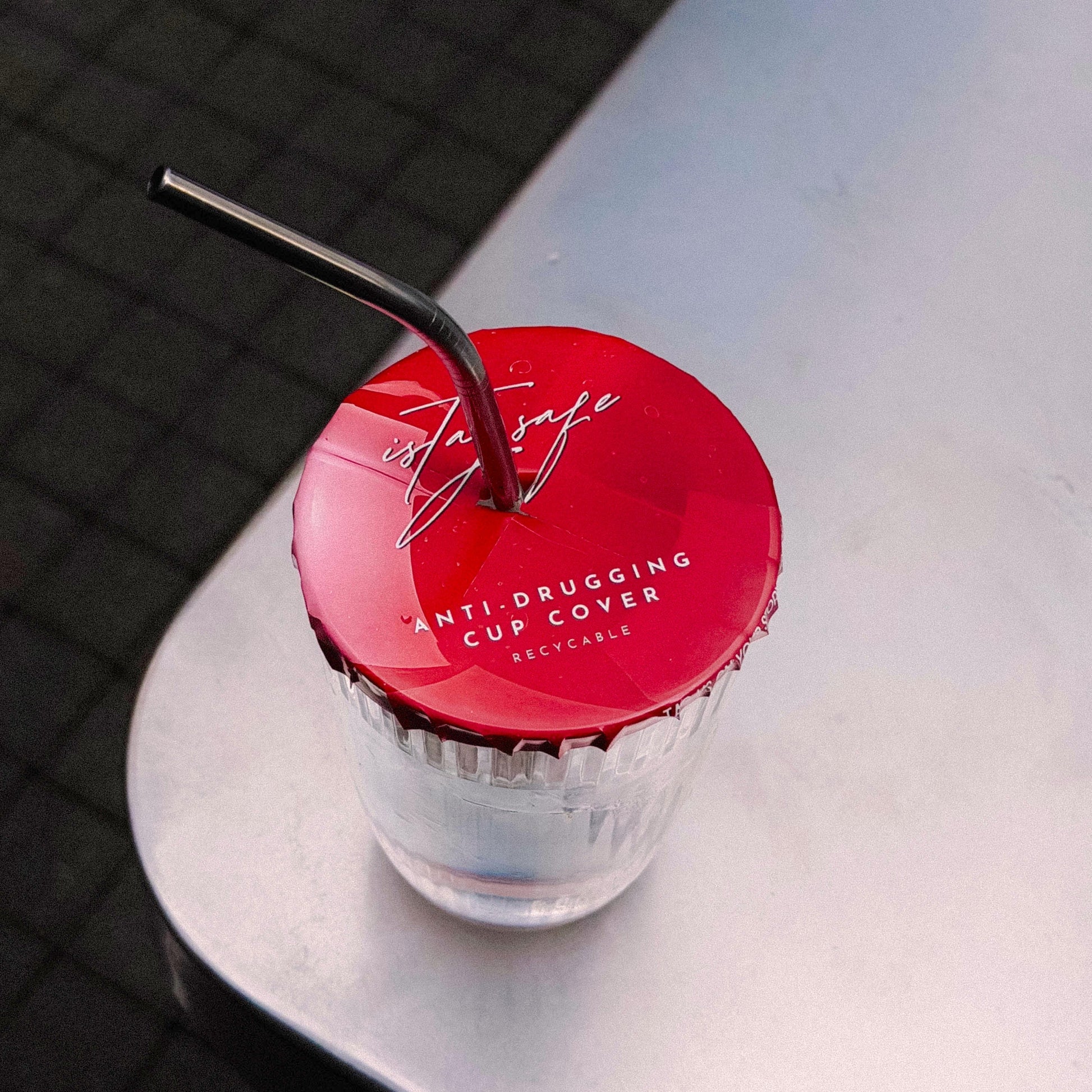 Clear cup with red ISTAYSAFE cup cover and straw on a reflective surface