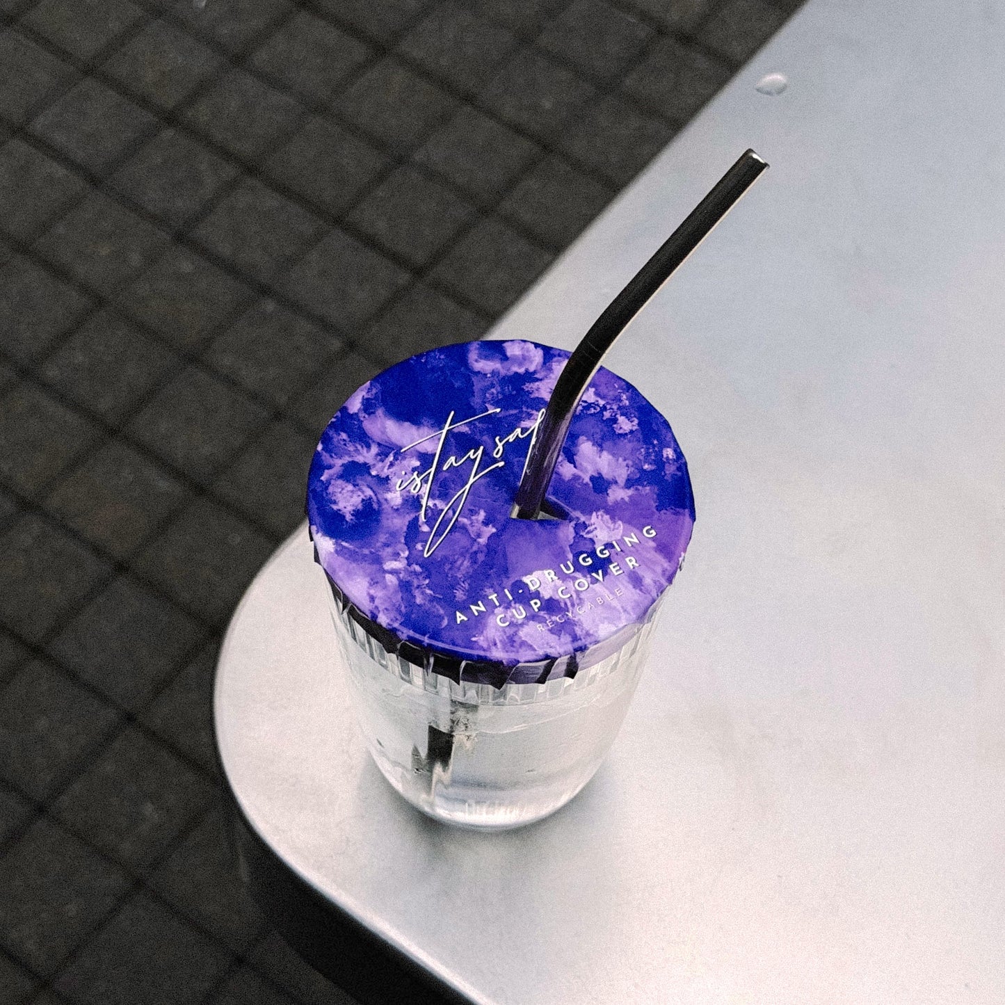 Clear glass with purple ISTAYSAFE cup cover and straw on a reflective surface