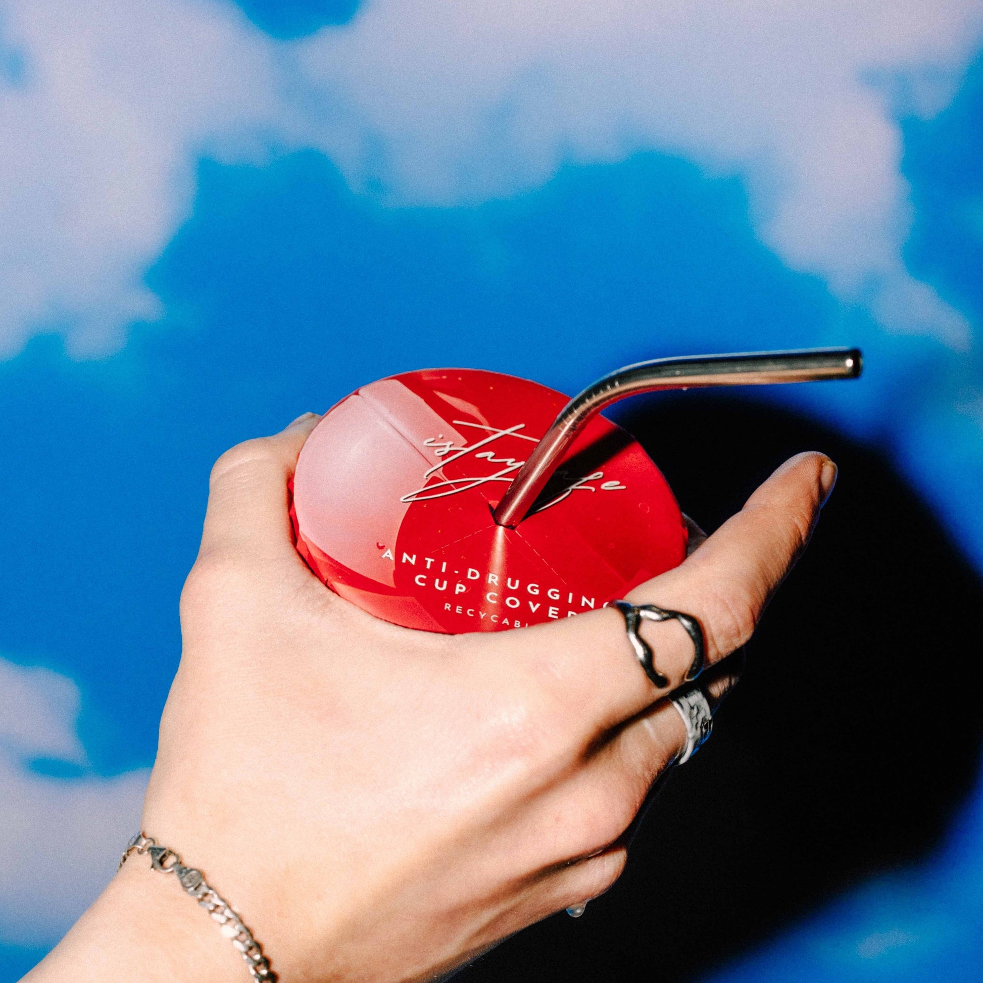 Hand holding a red ISTAYSAFE cup cover with a metal straw against a blue background