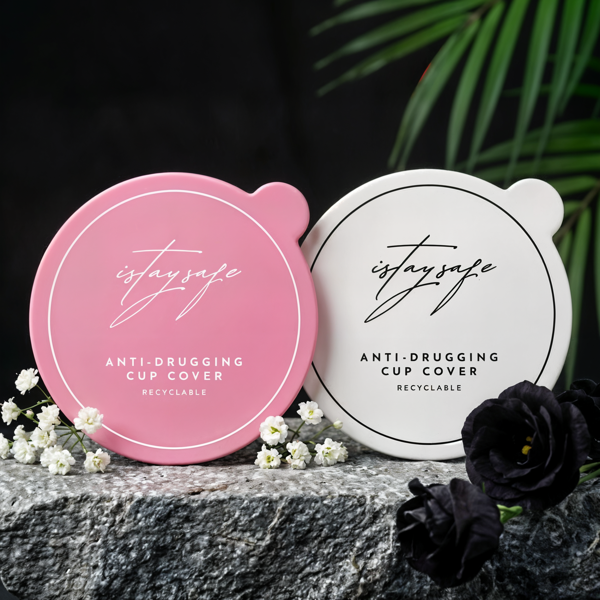 Two ISTAYSAFE anti-drugging cup covers, one pink and one white, on a stone surface with flowers and greenery.